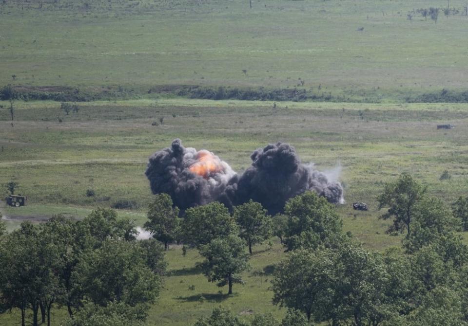 dark smoke rises from an explosion behind trees in an open field from an exploded munition