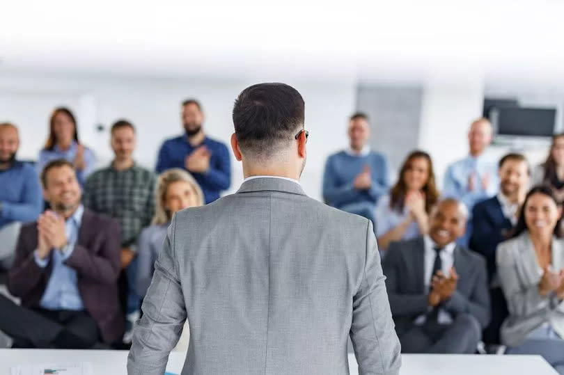 Rear view of a male public speaker talking on a business seminar in board room while his colleagues are applauding him. Copy space.