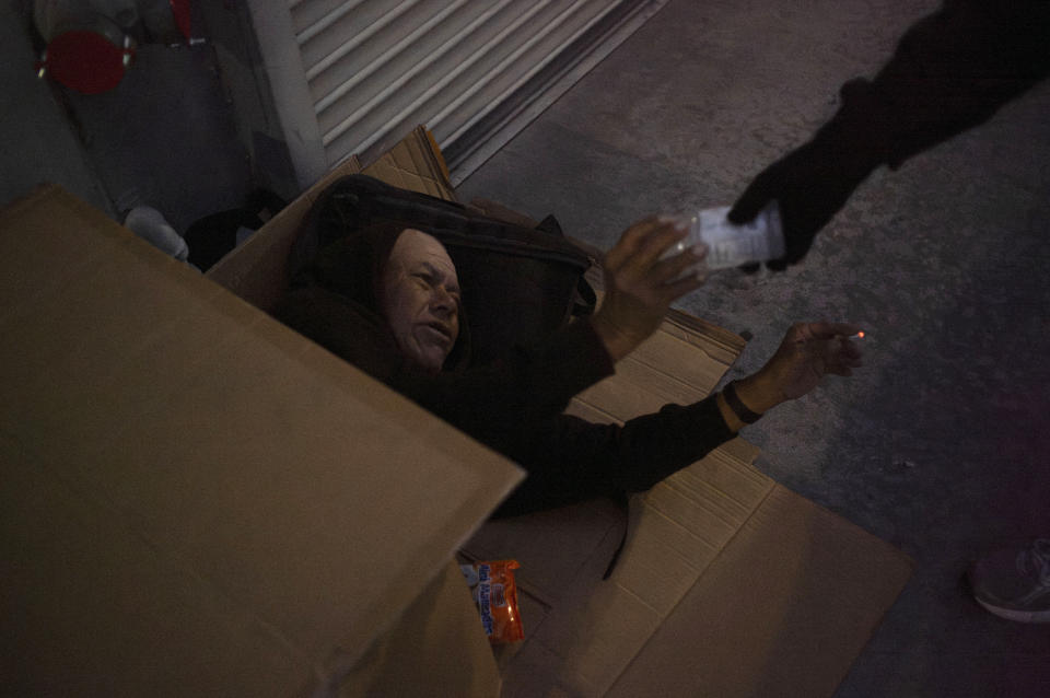 Francisco Sanchez, who has been homeless for nearly 30 years, lies in a makeshift cardboard shelter as he receives a bottle of sports drink from a passerby in Los Angeles, on Wednesday, Oct. 25, 2023. There is no single reason why Los Angeles became a magnet for homelessness. Two contributing factors: Soaring housing prices and rents punish those with marginal incomes, and a long string of court decisions made it difficult for officials to clear encampments or relocate homeless people from parks and other public spaces.(AP Photo/Jae C. Hong)