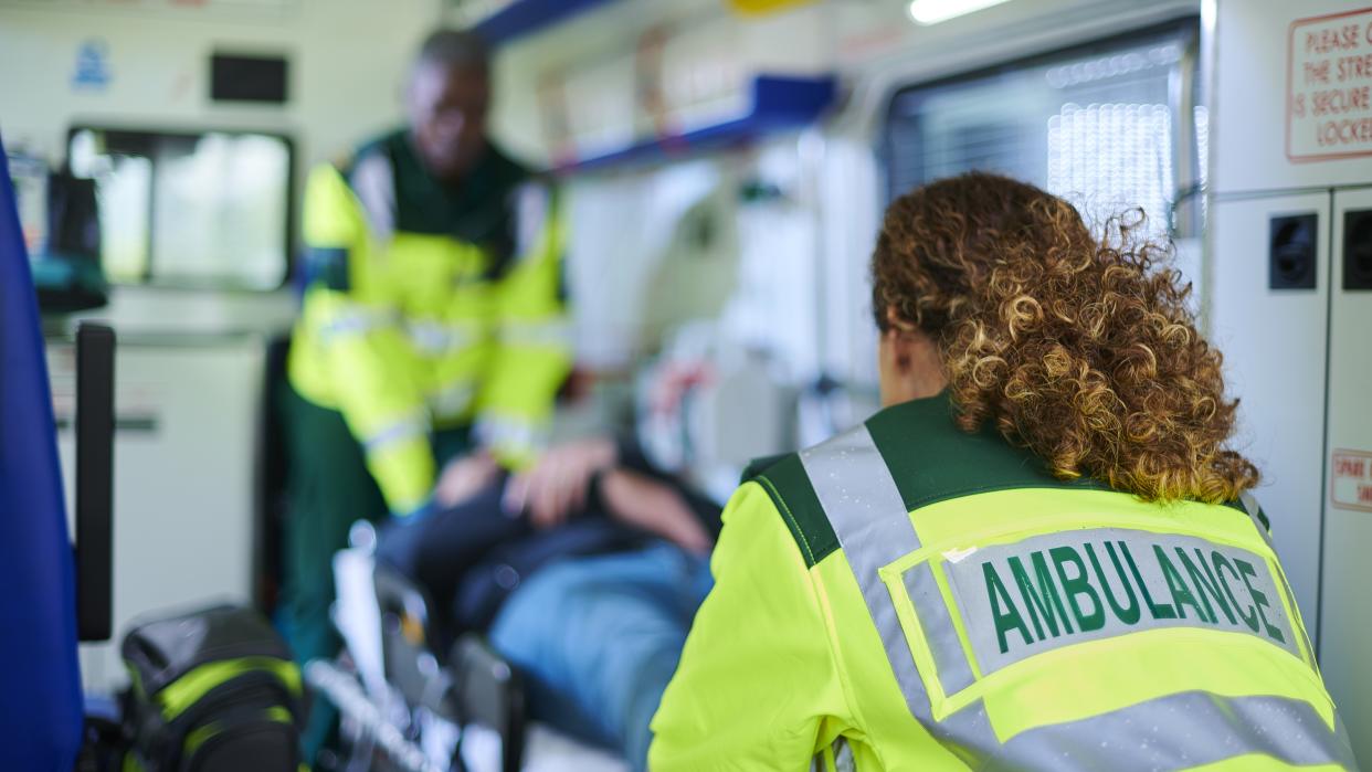  Two medical providers in an ambulance wearing bright uniforms and preparing to slide a man on a stretcher out of the vehicle. 