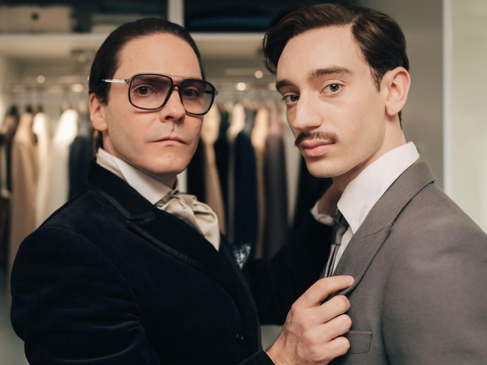 Daniel Brühl as Karl Lagerfeld wearing sunglasses and a dark velvet jacket, and Théodore Pellerin as Jacques de Bascher, a man with a mustache wearing a gray suit