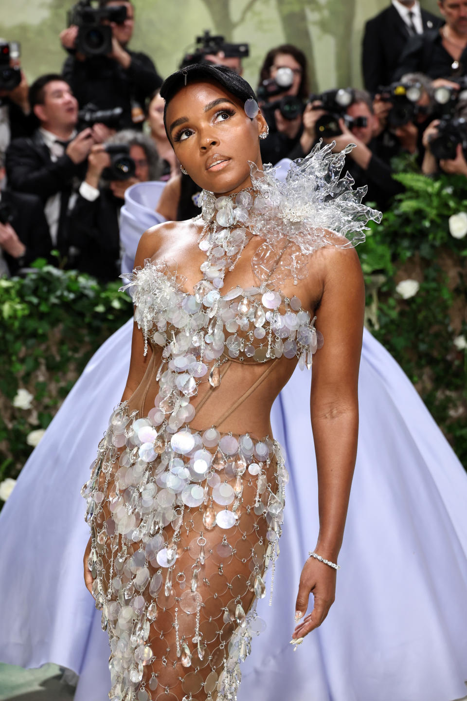 Janelle Monáe wearing a sheer, intricate dress with floral and crystal embellishments on a red carpet, surrounded by photographers