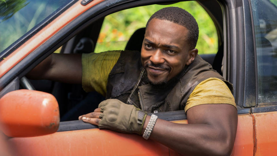 Anthony Mackie smiles while sitting in a car, wearing a brown vest, yellow shirt, and fingerless gloves