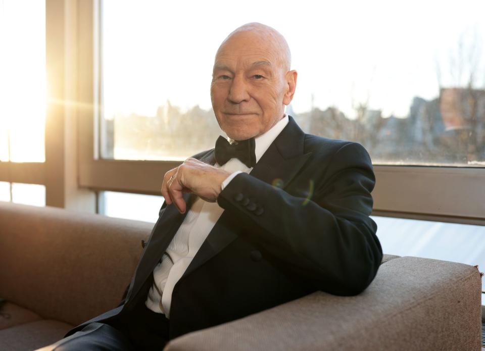 patrick stewart sits on a sofa with his left arm across him, and the sun shining through the window behind him