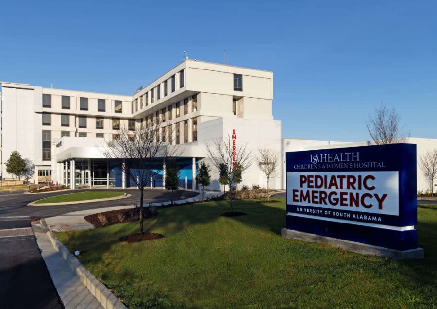Photo of a white hospital building. The front side has windows and it wraps around in an L shape. On the front of the building is a vertical sign that reads EMERGENCY in big, red letters. In the foreground of the photo there is a sign that reads "USA Health Pediatric Emergency"