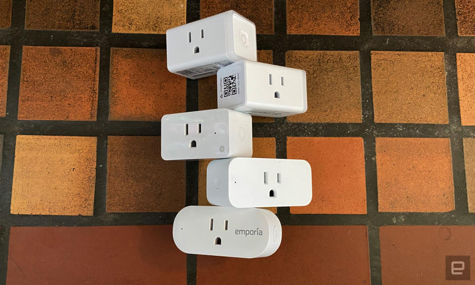 Five smart plugs from TP-Link, Amazon, Emporia and GE are stacked on a yellow, orange and brown tiled surface.  