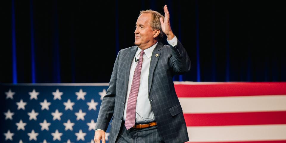 Texas Attorney General Ken Paxton waves in a suit in front of an oversized American flag