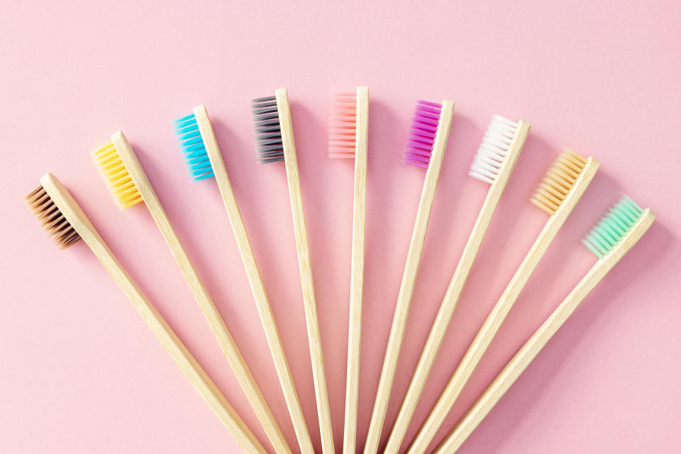 Nine bamboo toothbrushes arranged in a semi-circle on a flat surface. Each toothbrush has bristles of different colors