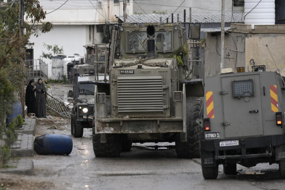 Palestinian women watch Israeli military vehicles, including a bulldozer, in Aqbat Jabr camp, southwest of the city of Jericho on Saturday, Feb. 4, 2023, during a search for the Palestinian suspects behind a shooting attack at a restaurant in a nearby settlement last week. The Israeli army raided the refugee camp near the Palestinian city of Jericho on Saturday, besieging houses it said were being used as hideouts for Palestinian attackers and shooting at residents who opened fire. (AP Photo/ Majdi Mohammed)