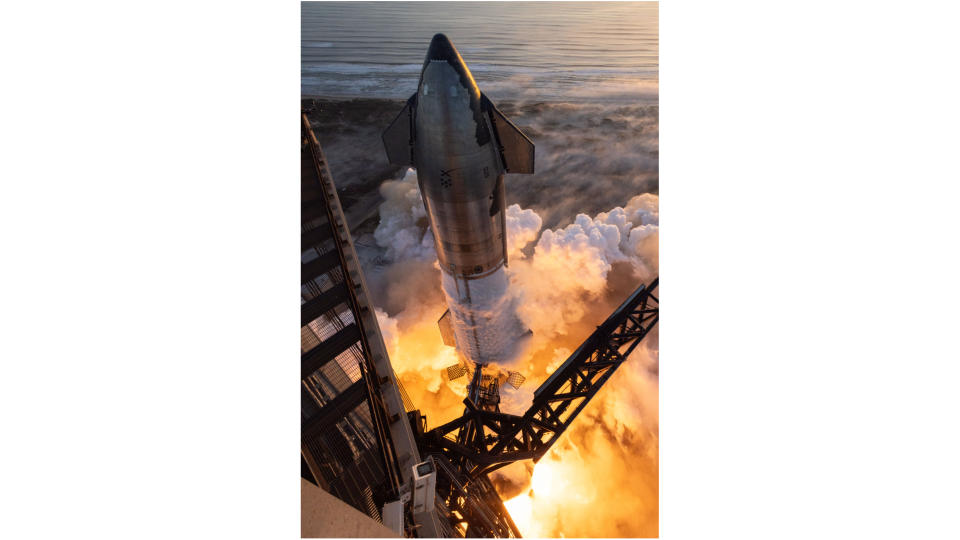 view looking down at a huge silver rocket on the launch pad, with flames billowing from its base.