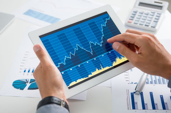 Two jacket-clad hands holding a tablet above a table full of charts and calculators. The right hand points to a chart on the screen, showing growth.