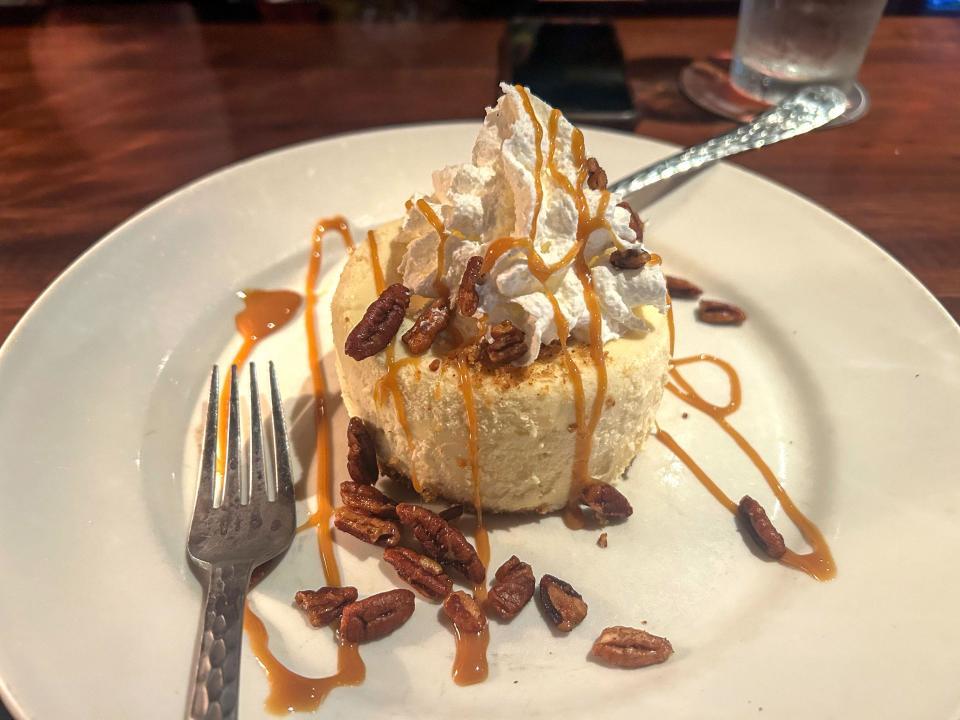 A small cheesecake topped with whipped cream, a caramel drizzle, and pecans. The cheesecake is served on a white plate with a fork.
