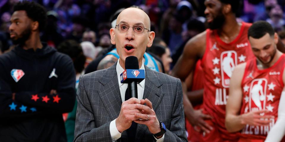 Adam Silver speaks into a microphone while players stand behind him at the All-Star Game.