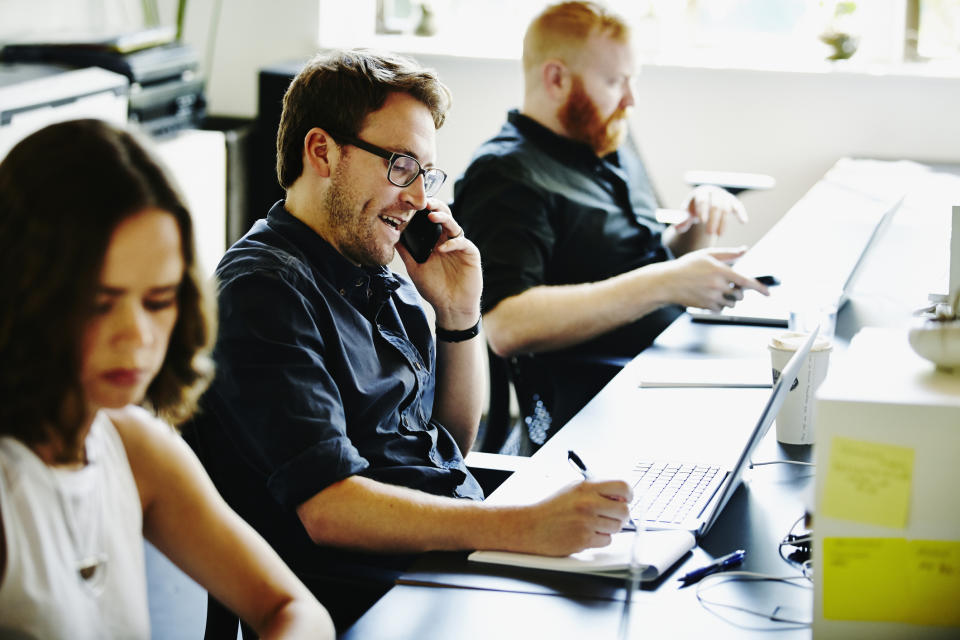 Three individuals working at a desk with laptops and phones, with the middle person smiling and talking on the phone