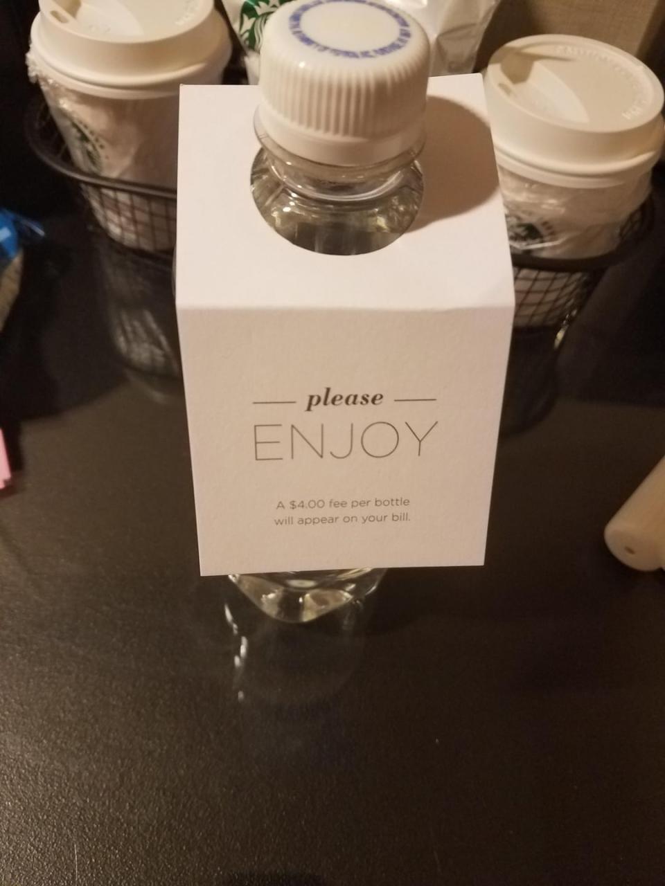 A hotel water bottle with a tag reading "please ENJOY - A $4.00 fee per bottle will appear on your bill."