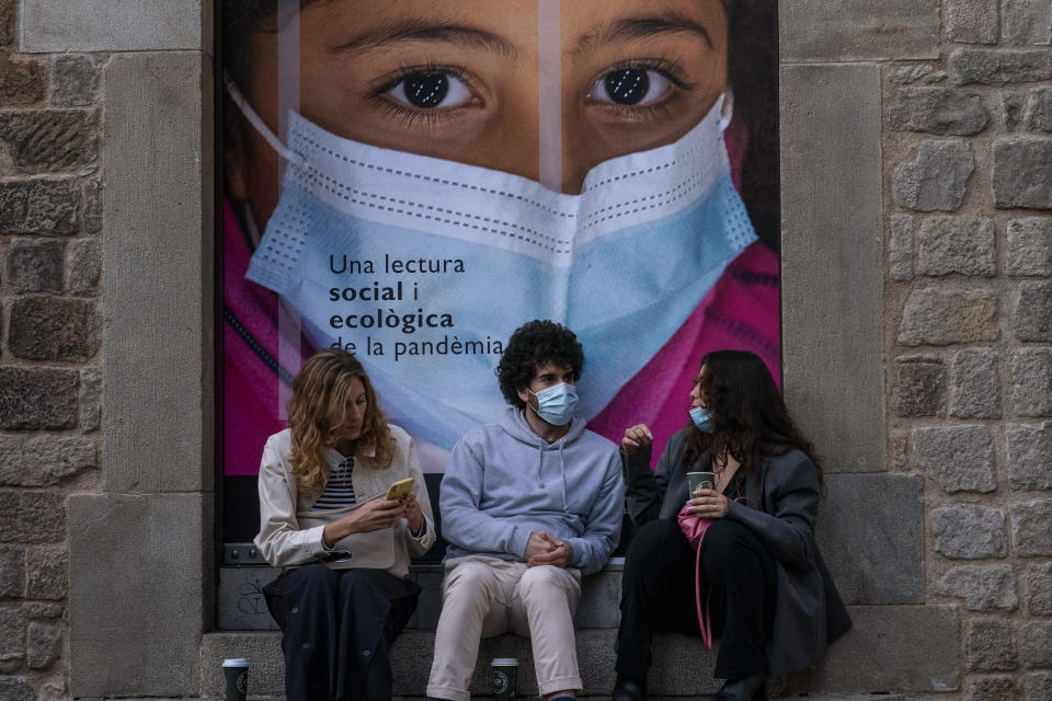 People, some of them wearing face masks, sit next to an advertising poster in downtown Barcelona, Spain, Friday, April 23, 2021. (AP Photo/Emilio Morenatti)