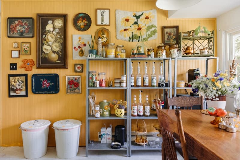 Vintage floral paintings and prints surround food storage shelves in yellow kitchen.