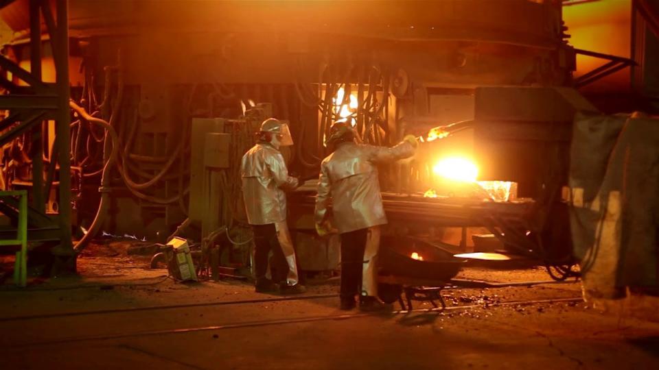 Two workers wearing heat-resistant clothing near a blast furnace.