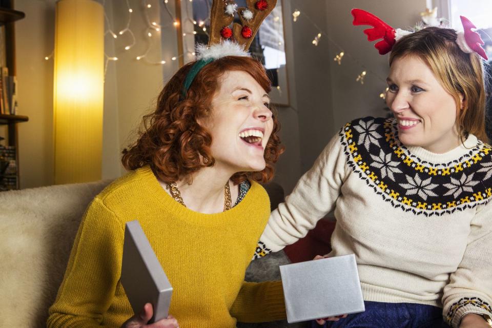 redheaded woman smiling when sitting on a couch opening a christmas present in a silver box there is a woman in a sweater beside her smiling she is wearing pink and red antlers on top of her head
