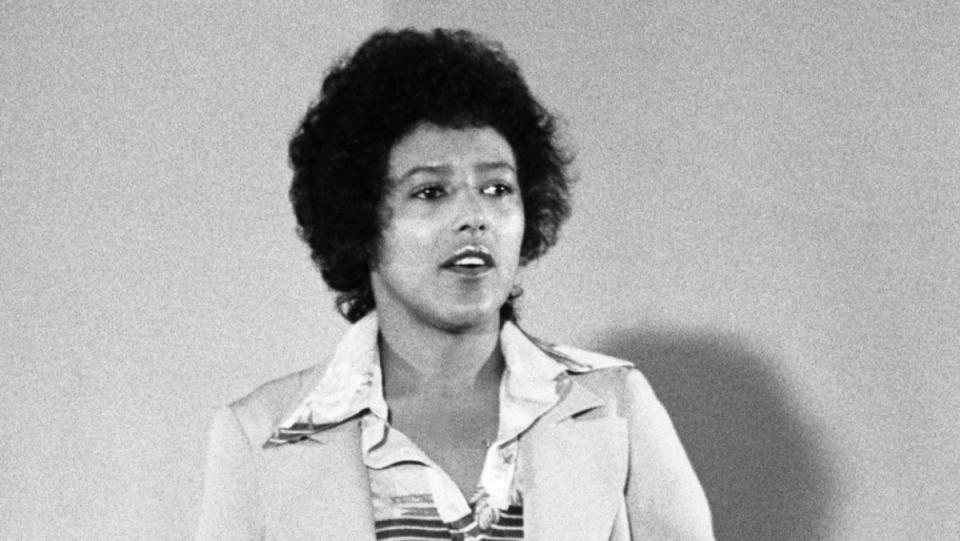 a Black and white photo of a woman named Elaine Brown wearing a soft curly black Afro and a pantsuit with a silk shirt 