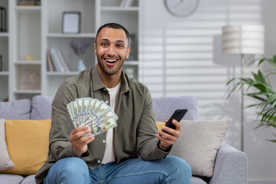 Portrait of a young happy Latin American man sitting, holding cash dollars and a phone in his hands, pointing to the camera, smiling, happy.