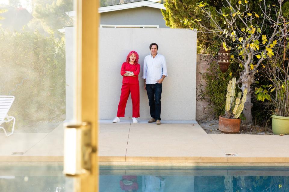 A woman in red and a man in a white shirt and black pants stand in front of a pool in his back yard.