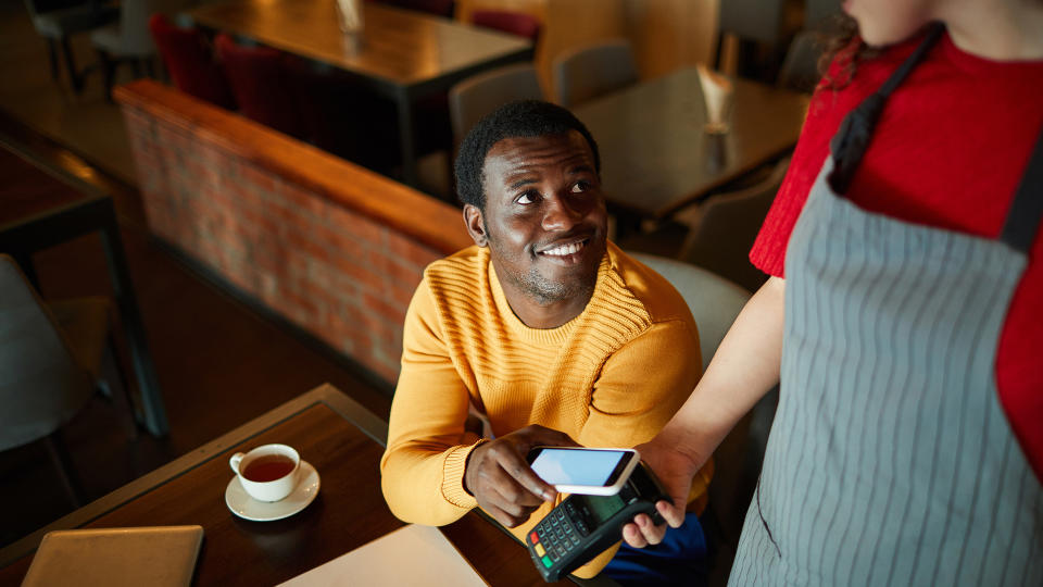 Young smiling man sitting by table and looking at waitress while paying for his order through smartphone.