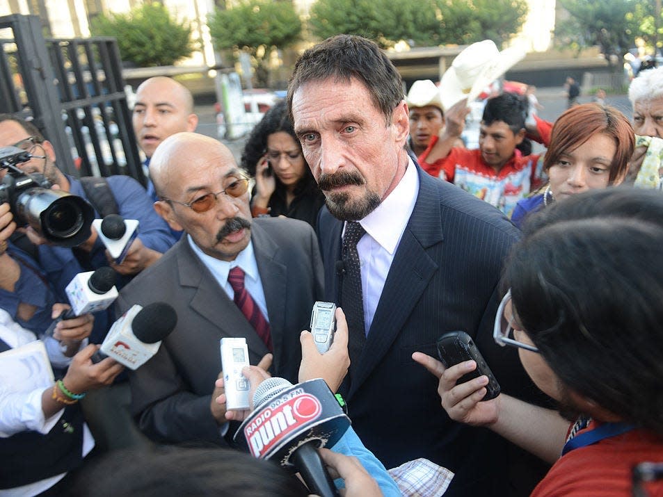 John McAfee, with dark hair and a dark mustache and beard, stand amid reporters holding microphones.