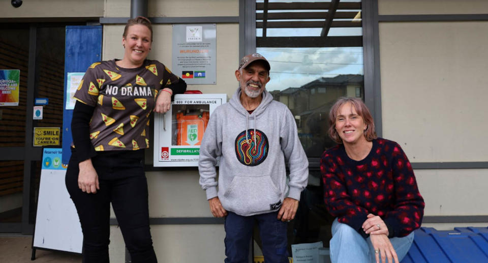 A photo of three locals posed around a defibrillator outside Reservoir Neighbourhood House.