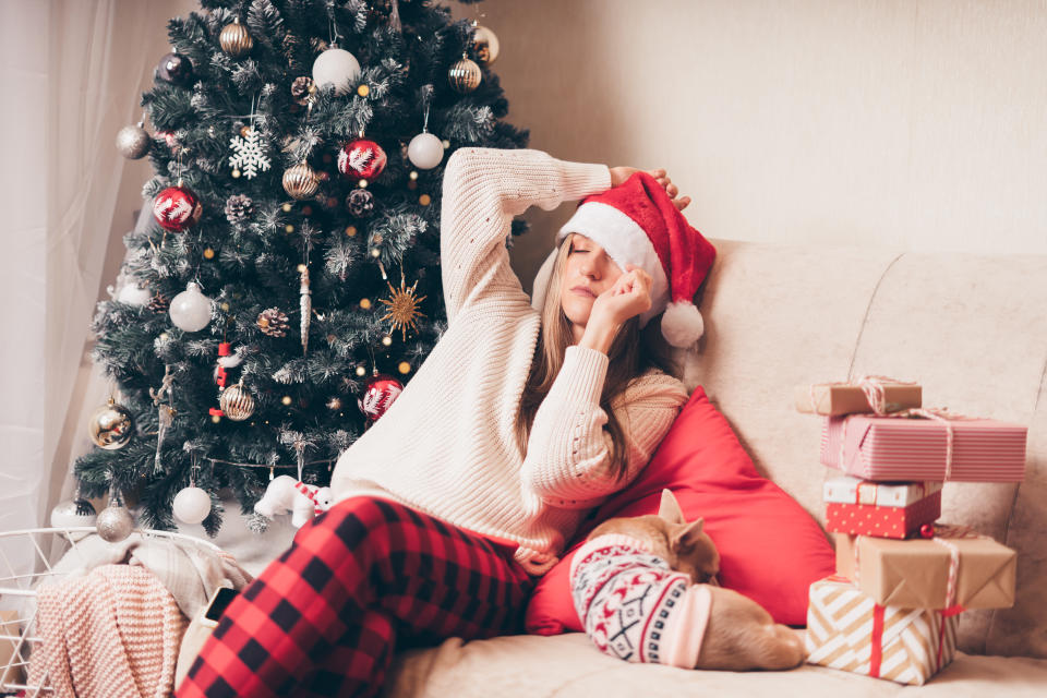 Woman falling asleep and tired of gift wrapping with puppy dog on a couch in the living room with Christmas tree at home