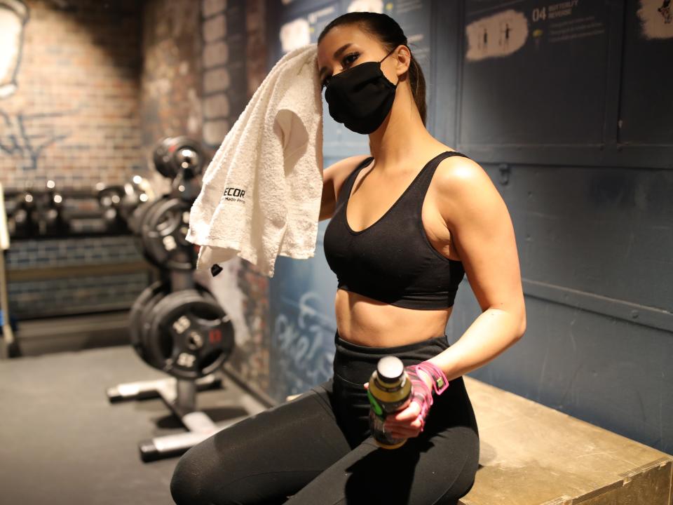 A woman in a fitness room wears a protective face mask, wiping her face with a towel.