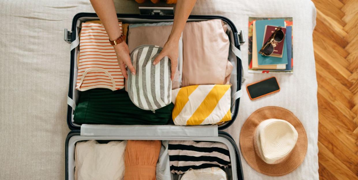 shot from above of woman packing things in her suitcase on the bed she is holding and putting a grey and white stripped cosmetics bag in there is her mobile phone, books and sunglasses on the bed