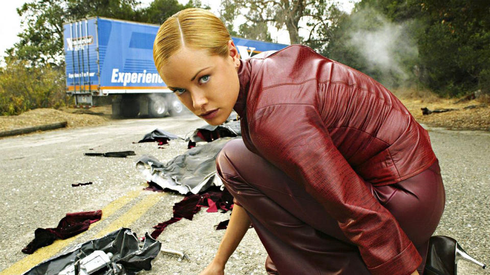 Still from a Terminator movie. An immaculate, blonde-haired cyborg wearing a red leather jacket and red leather pants is crouching down on the road. Behind here there is a large truck smoking and bits of truck debris everywhere.