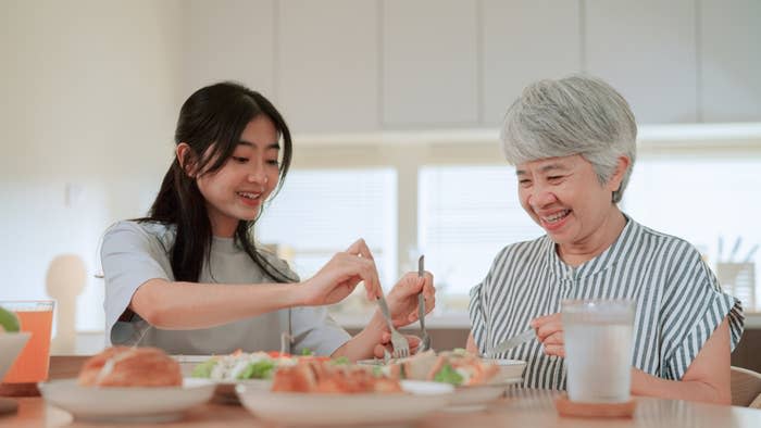 A young woman and an older woman are enjoying a meal together at a dining table. They are smiling and appear to be sharing food