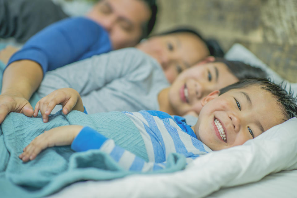 An East Asian family of four sleep in the same bed, with their two children beaming at the camera as their parents spoon them