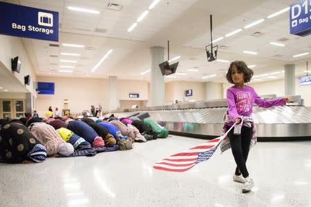 A young girl dances with an American flag in baggage claim while women pray behind her during a protest against the travel ban imposed by U.S. President Donald Trump's executive order, at Dallas/Fort Worth International Airport in Dallas, Texas, U.S. January 29, 2017. REUTERS/Laura Buckman