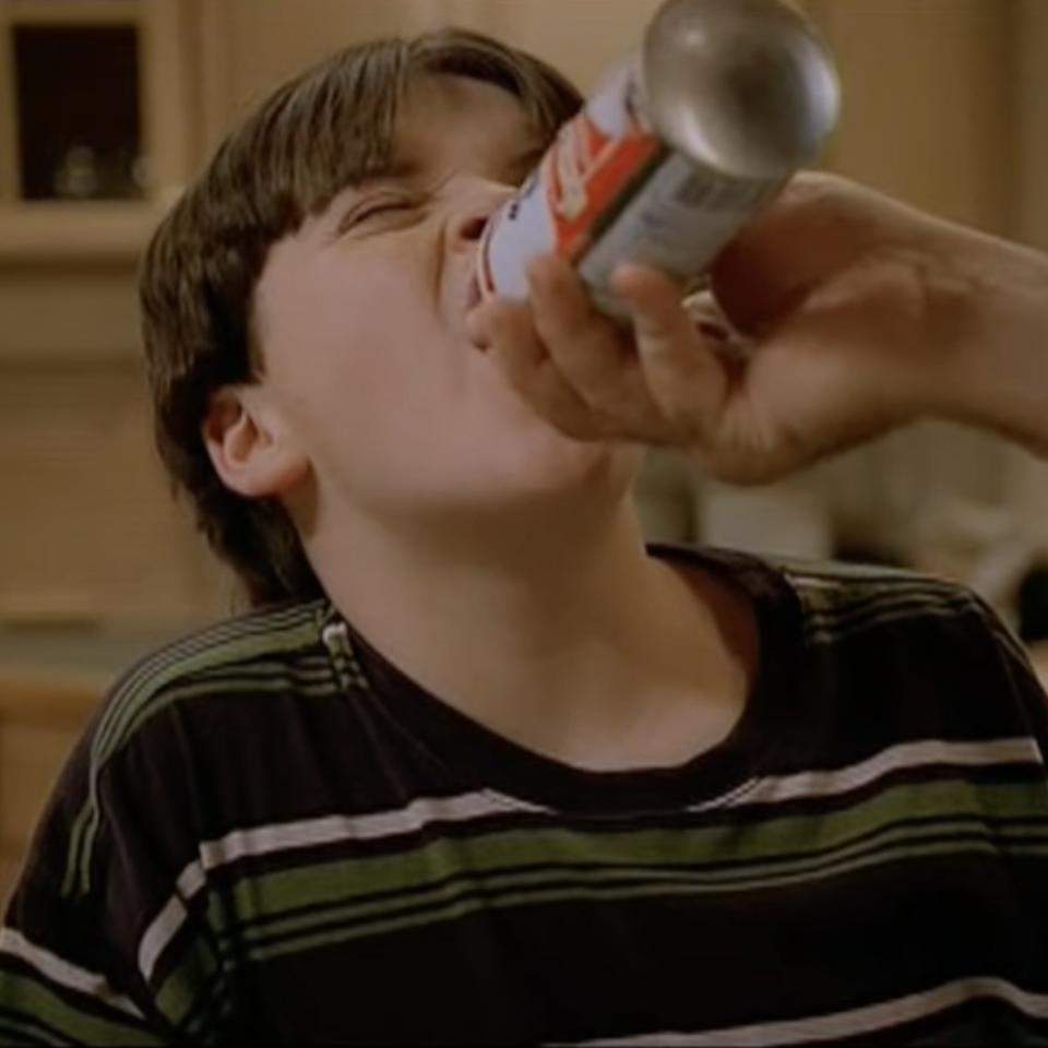 A person, hand and arm not visible, pours an aerosol can directly into the open mouth of a young boy with straight dark hair in a striped shirt