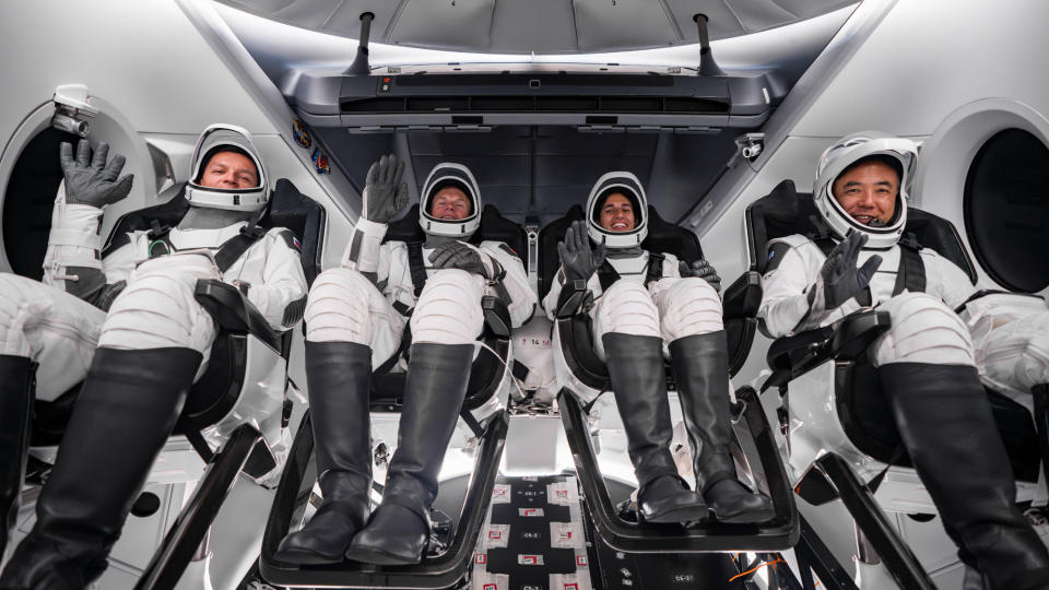 four astronauts in white spacesuits smile while seated inside a spacecraft capsule