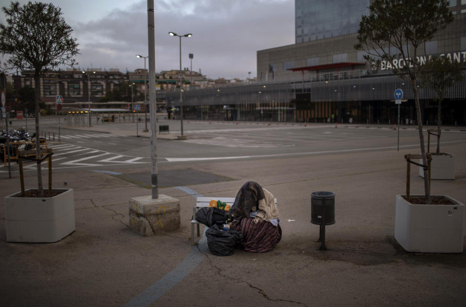 In this Sunday, March 22, 2020 photo, a man from sub-Saharan Africa covers himself with clothes and blankets as he sits on a bench at an empty car park outside the train station in Barcelona, Spain. (AP Photo/Emilio Morenatti)