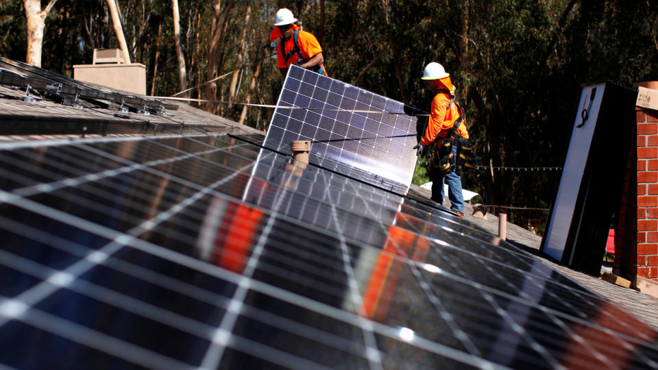 Two workers add a solar panel to a roof that already has several panels in place.