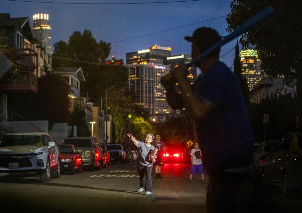 A teenage girl pitches a ball against the city skyline at dusk.