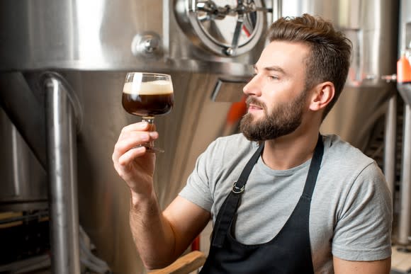 Brewer looking at a snifter of dark beer, with brew tanks in the background