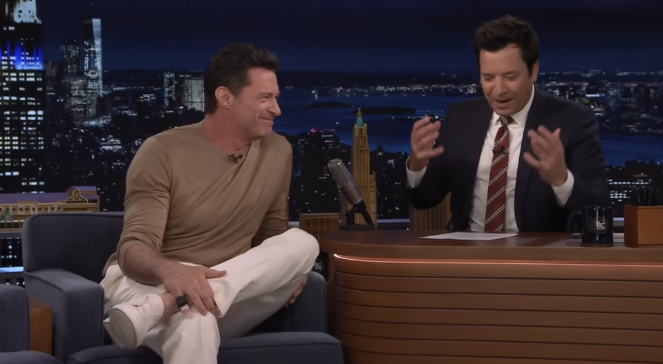 Hugh Jackman sits on a talk show set with his legs crossed, chatting with a gesturing Jimmy Fallon during a nighttime show