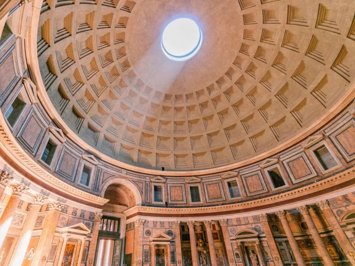 The dome of the Pantheon is shown with light beaming through its center. This intricate concrete structure is made up of beveled squares that line the whole surface of the dome.