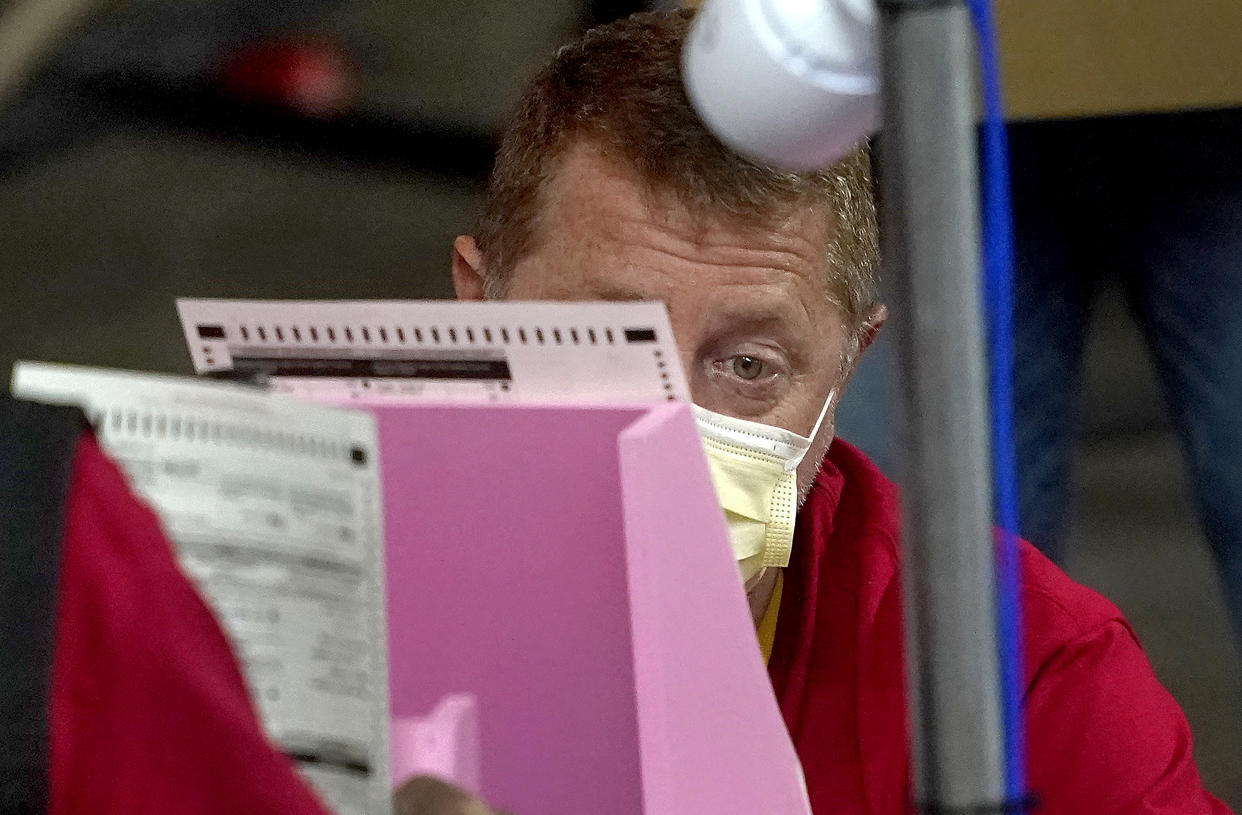 A worker in a red shirt and yellow face mask closely scrutinizes a ballot propped against a pink stand.