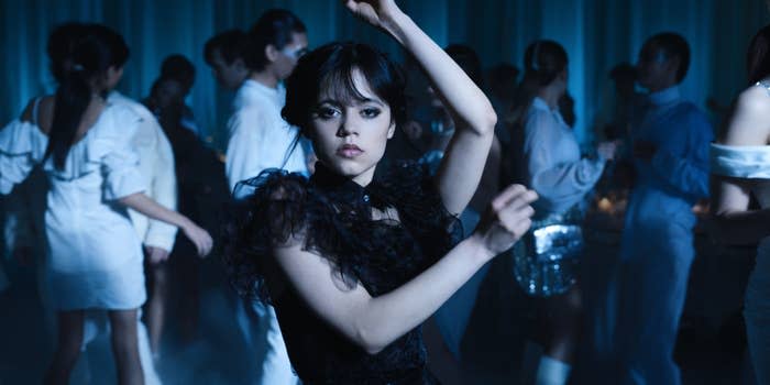 Jenna Ortega, in a gothic-style dress, dances amidst a crowd at a dimly lit event in a scene from "Wednesday"