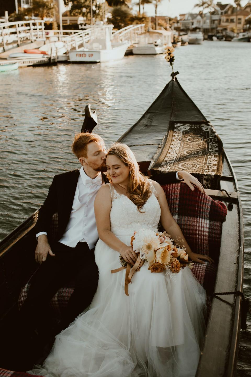 A groom kisses his bride's head as they sit in their wedding attire on a gondola.