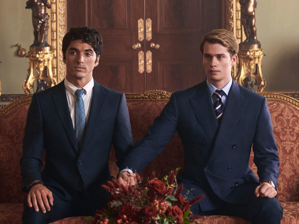 alex and henry in red white and royal blue, holding hands and sitting on an ornate chaise in a lavish, royal room. the king and henry's brother sit in front of them