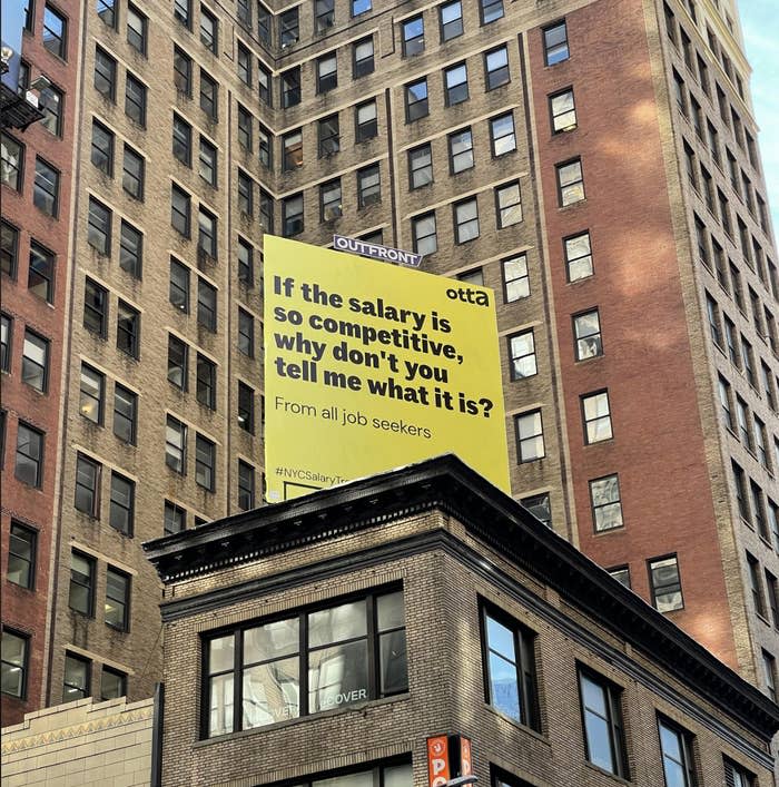 Billboard above city street questioning why competitive salaries aren't disclosed, engaging job seekers. #NYCSalary #InDoors