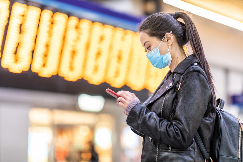 Chinese woman wearing face mask at train station to protect from smog and virus - young asian woman looking at her smartphone with departure arrivals board behind - health and travel concepts